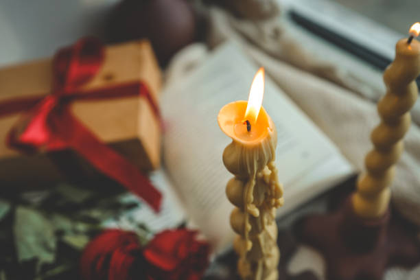 Close-up of a twisted candle with a burning flame, wax drips creating a textured surface, blurred background featuring a red ribbon gift box, roses, and an open book for a cozy ambiance