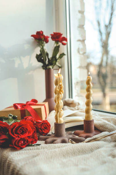 Two twisted candles burning in modern holders by a windowsill, accompanied by a red ribbon gift box, red roses, and a vase with roses, creating a cozy ambiance with natural light