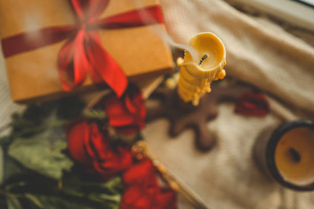 Twisted extinguished candle with soft swirling smoke in a cozy setting, accompanied by a kraft gift box tied with a red ribbon, red roses, and blurred decor creating a warm, romantic ambiance.