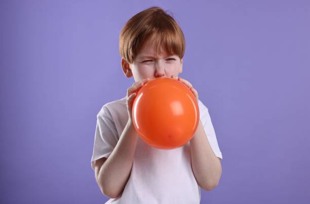 istock Boy inflating orange balloon on violet background 2193066470