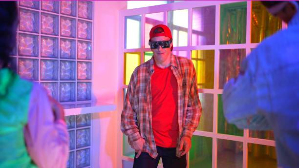 Caucasian dancer dancing while diverse friend looking at footstep in neon light at night club. Back view of multiethnic hipster moving to freestyle music with happy man looking at camera. Regalement.