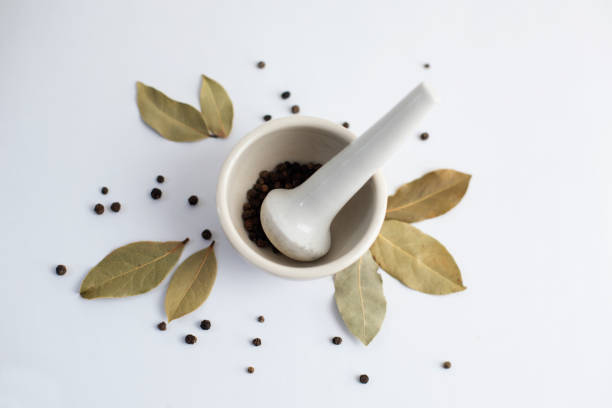 White mortar with black pepper and bay leaves on a white background.