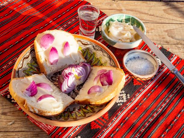 Traditional Bulgarian breakfast with bread, lard, and red onions on a colorful tablecloth, accompanied by salt and a shot glass.