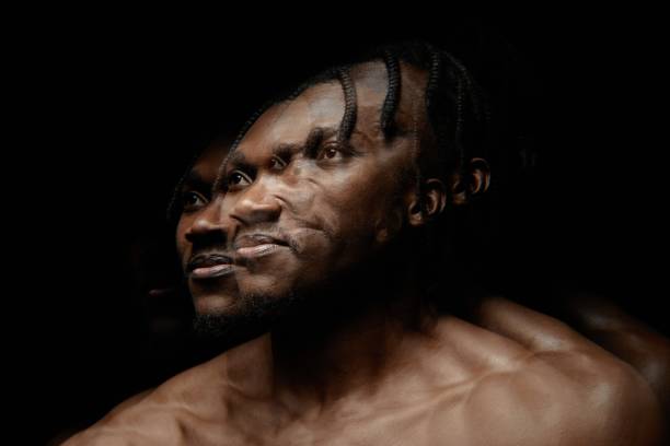 Image with double exposure effect of African American man looking up while posing shirtless in contrast light against black background in studio, copy space