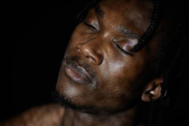 High angle shot of African American man with eyes closed looking up at top light drown in inner thoughts while standing in dark studio, copy space