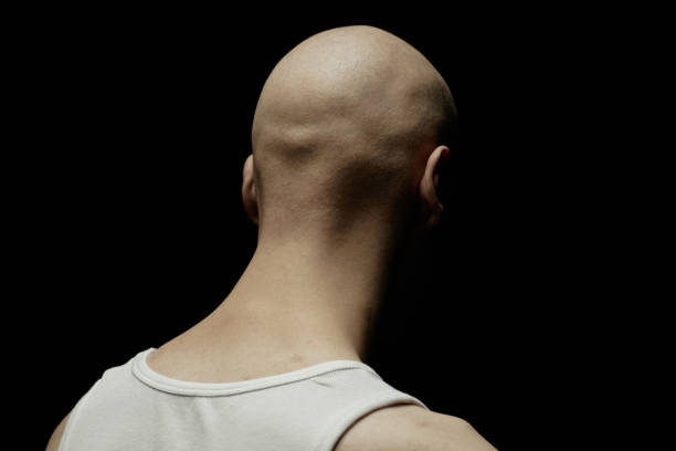 Rear view of pale bald man with face hidden by shadow looking deep into darkness in feeling of despair while standing in dimly lit studio, clean silhouette lines, copy space