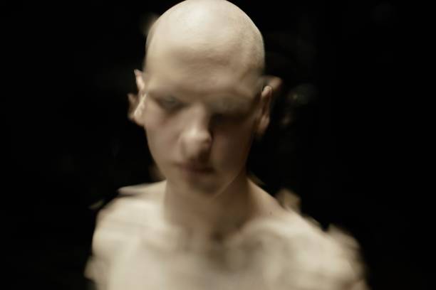 Distorted image of pale bald man staring with scary look at camera through glass with running down water against black background in studio, mental, health, disorder concept, copy space