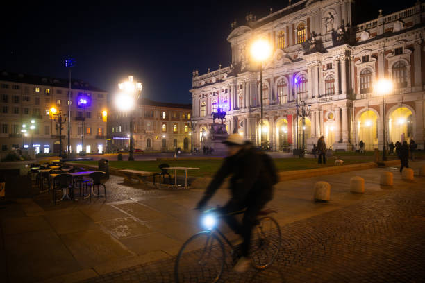 A lively evening scene in Turin, Italy, showcasing a cyclist passing through a beautifully illuminated square. The square features cobblestone paths, outdoor seating, and street lamps, creating a vibrant yet cozy atmosphere. Perfect depiction of urban life blending with historic charm in this iconic Italian city.Location: Carlo Alberto Square, Turin, Italy