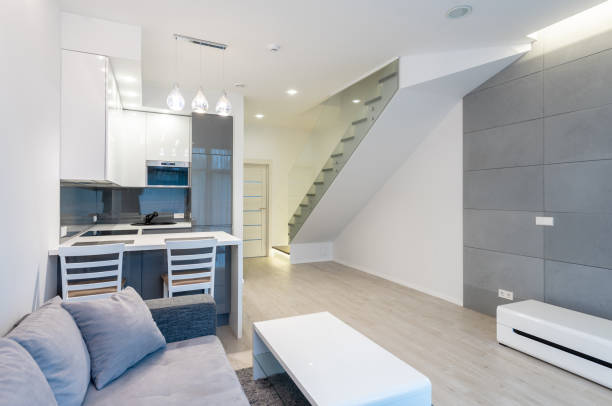 A bright, modern apartment showcasing a seamlessly integrated kitchen and living space. Light grey and white tones create a clean, spacious feel. A staircase with glass balustrade. Home Interior