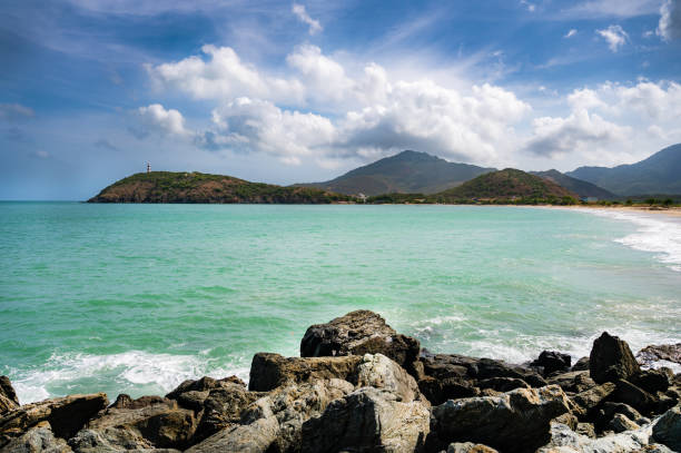 Lighthouse is on a hill overlooking the ocean. The water is calm and blue. The sky is clear and blue