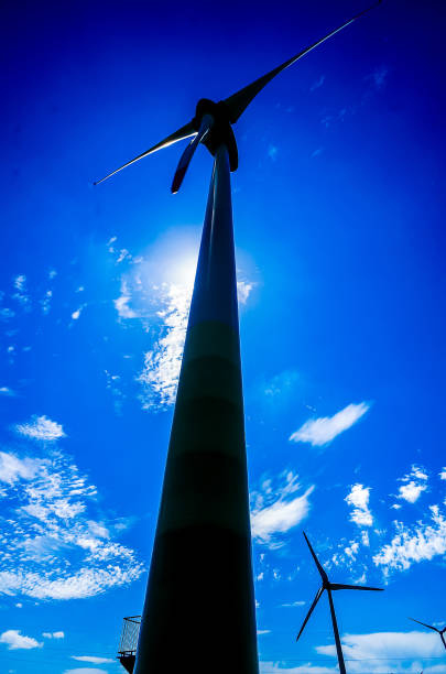 A wind turbine is standing tall in the sky above a blue sky. The sun is shining brightly on the turbine, making it look like a giant light post. The scene is peaceful and serene
