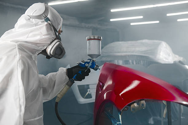 istock Car paint worker spraying red paint to car body element using spray gun in vehicle workshop chamber. Complex restoration of automobile body. 2193109123