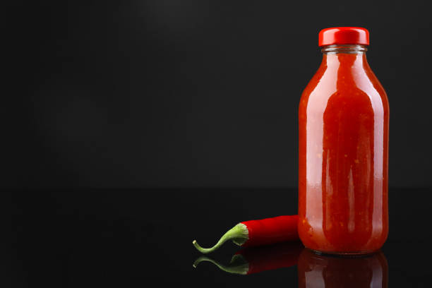 Spicy chili sauce in bottle and pepper against dark background, space for text