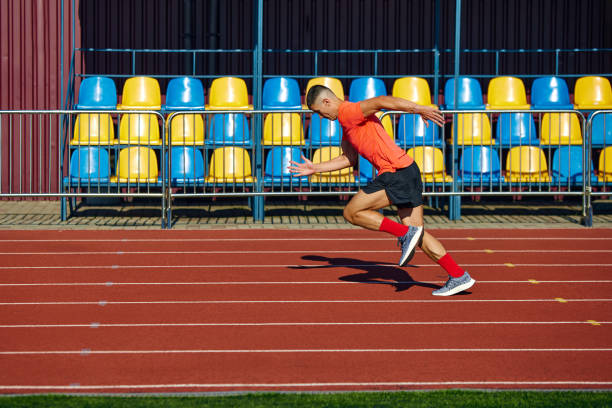 istock Man, sprinter in bright red shirt propels himself down track, his form sharp against backdrop of colorful, empty stadium seats. 2193117038