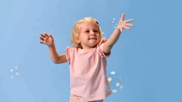 istock Happy little child, smiling girl clapping hands happily while catching soap bubbles wearing pink clothes with curly hair against blue studio background. 2193117122