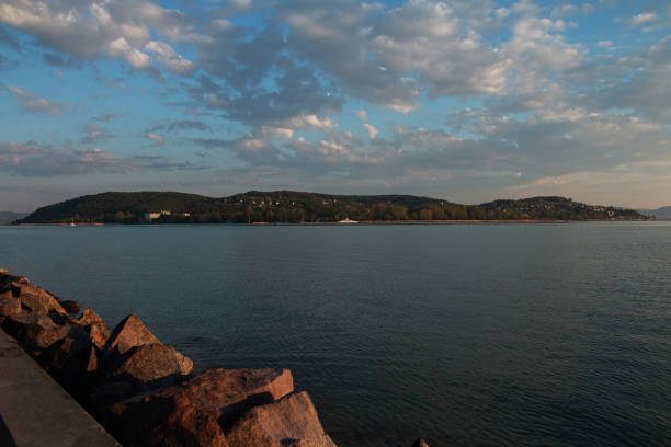 The early morning light gently illuminates the Balaton Lake, as seen from the Szántód ferry port. The blue sky, adorned with soft white clouds, stretches over the tranquil waters and the distant Tihany Peninsula.