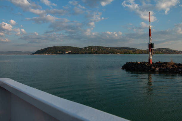 The early morning light gently illuminates the Balaton Lake, as seen from the Szántód ferry port. The blue sky, adorned with soft white clouds, stretches over the tranquil waters and the distant Tihany Peninsula.