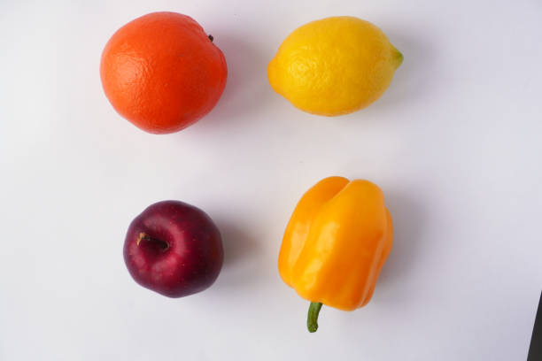 A vibrant arrangement of an orange, a lemon, a yellow bell pepper, and a red apple placed on a clean white background, highlighting their natural colors and textures in a minimalistic style.