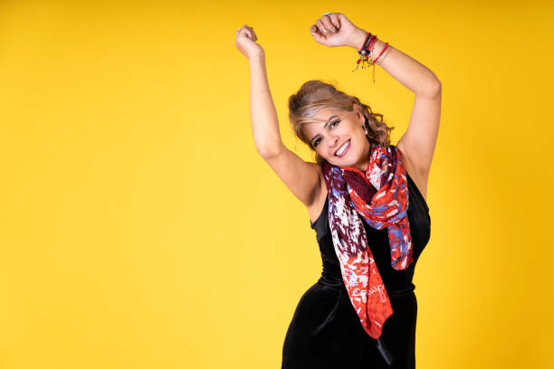 Portrait of a cheerful mature woman dancing with arms raised against a vibrant yellow background