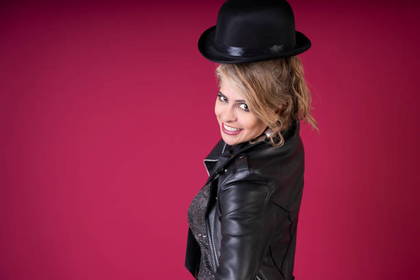 Smiling female singer wearing a bowler hat and leather jacket, posing on a pink background