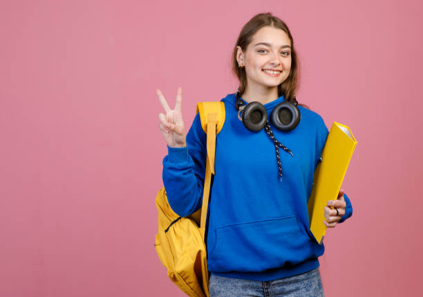 Front view of brunette girl standing, smiling, showing peace, greeting. Pretty schoolgirl with yellow rucksack and folder looking at camera, happy. Concept of education and youth.