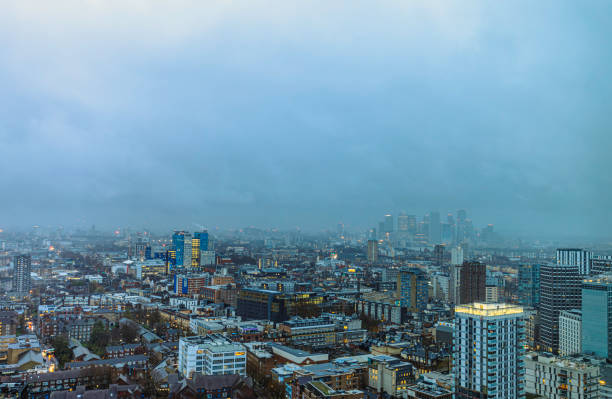 A panoramic view of London's sprawling cityscape under a moody, overcast sky. The distant skyscrapers of Canary Wharf emerge through the rain and mist, while the foreground balcony adds depth to the urban landscape. A scene that captures the essence of London's metropolitan atmosphere during a rainy evening.