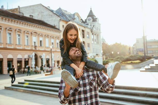 Father carrying his daughter on shoulders, both smiling joyfully in bright urban setting during golden hour, while enjoying travel and city break