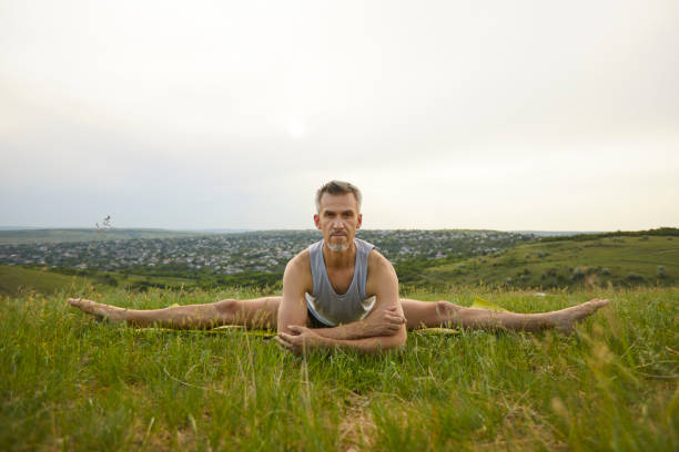 Serious middle aged male yogi doing Wide Angle Seated Forward Bend aka Upavistha Konasana during yoga workout in nature. Confident fit man looking at camera practicing straddle splits on green grass