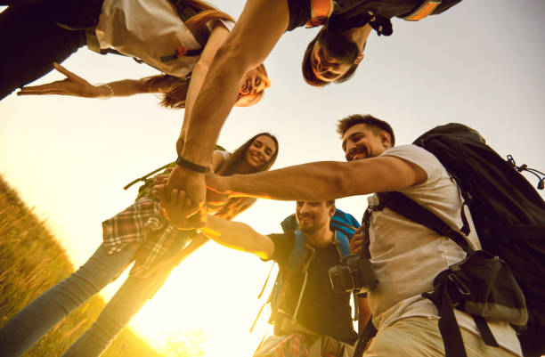 Low angle of team of tourists with rucksacks joining hands in field while hiking in countryside stock photo