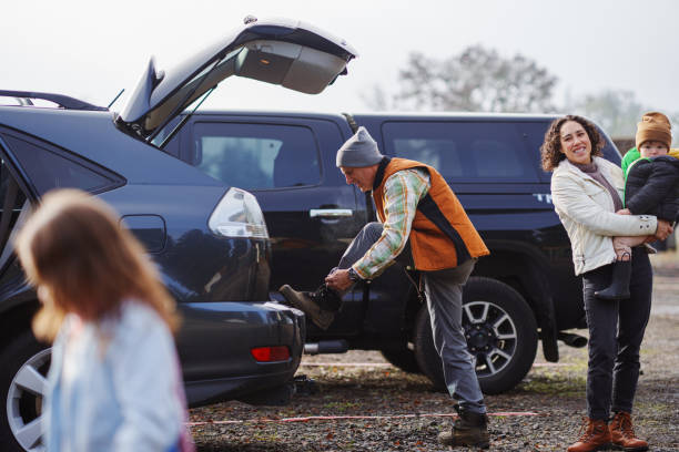 An vibrant and healthy senior man laces up his boots in the parking lot of a Christmas tree farm as his adult daughter and her young children wait so they can look for a tree together.