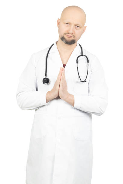 A bald male doctor with a beard is wearing a white lab coat over red scrubs and has a stethoscope around his neck. He is standing with his hands pressed together in a calm and composed gesture, conveying gratitude or a request. The plain white background emphasizes professionalism and focus