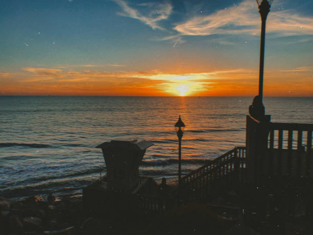 Stunning ocean sunset with vibrant orange and yellow hues, a lifeguard tower, and silhouetted figures on a wooden deck overlooking the serene waves. A peaceful coastal scene.