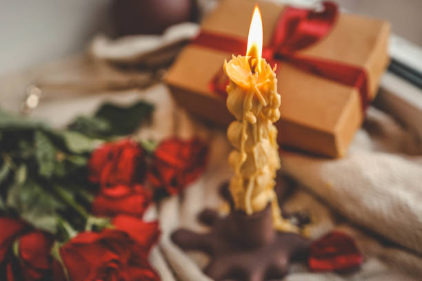 istock A cozy window-side scene featuring a lit beeswax candle with a unique shape, vibrant red roses, a gift box with a ribbon, and a soft textured fabric, creating a warm and serene ambiance 2193218103
