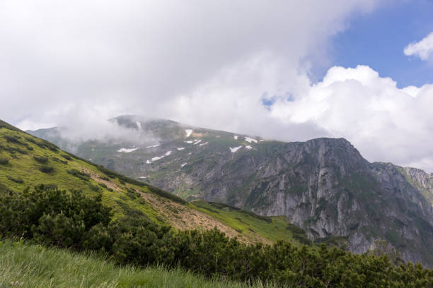 istock .Hilly terrain with green slopes interspersed with grass, bushes and rocks, with pointed mountain peaks partially covered by clouds in the background. 2193223555
