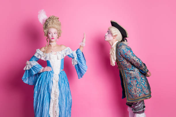 istock Elegant couple in costumes posing against a pink backdrop. The man leans forward, while the woman stands with a nonchalant expression 2193227615