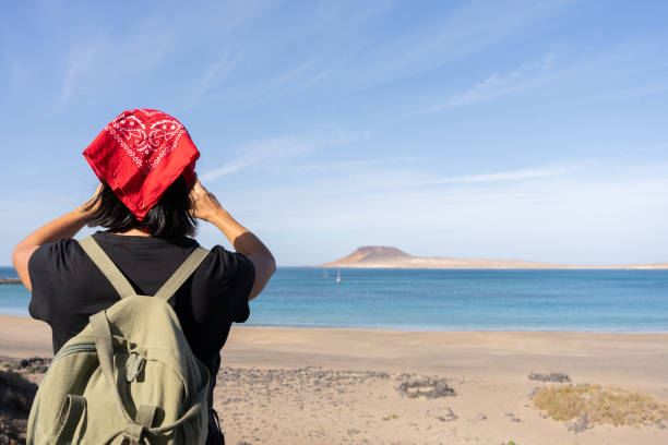 Female hiker fixing red bandana, gazing at Isla Graciosa landscape from Lanzarote volcanic terrain, Canary Islands, Spain