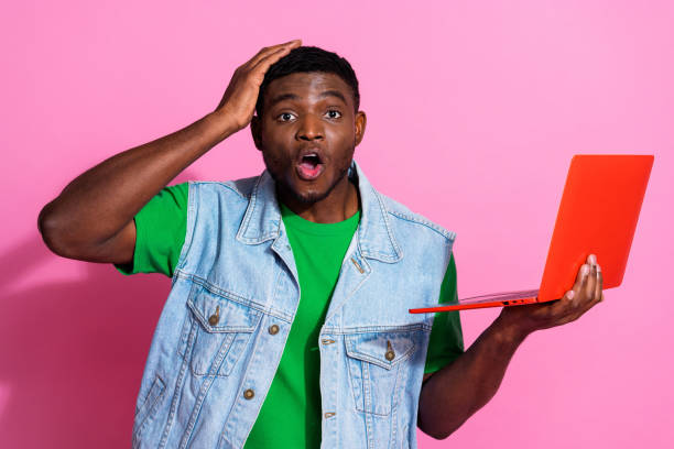 istock Young man with surprised expression holding a laptop against a vibrant pink background, showcasing a stylish casual look 2193229665