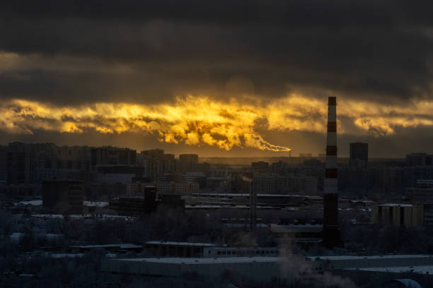 A captivating winter scene featuring smokestacks emitting smoke into the colorful backdrop of a vivid sunset. The snowy trees in the foreground and the urban skyline in the distance create a harmonious blend of industry, nature, and the warmth of fading daylight.