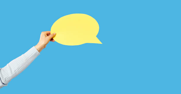 Human hand holding yellow paper mockup copyspace speech balloon, conversation cloud or message bubble against bright blue studio background. Cropped shot. Communication concept