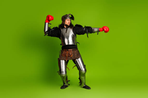 istock Young man dressed as a knight wearing boxing gloves and performing a dramatic stance against green background 2193243289