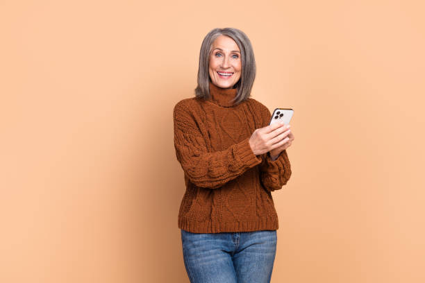 istock Elegant mature woman joyfully using a smartphone against a beige background, showcasing a trendy pullover and stylish casual outfit 2193247031