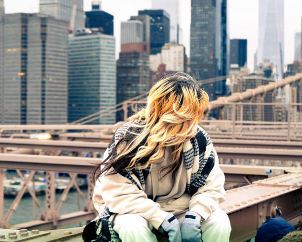 A woman with dyed hair sits on the ledge of the Brooklyn Bridge overlooking the Manhattan skyline.
