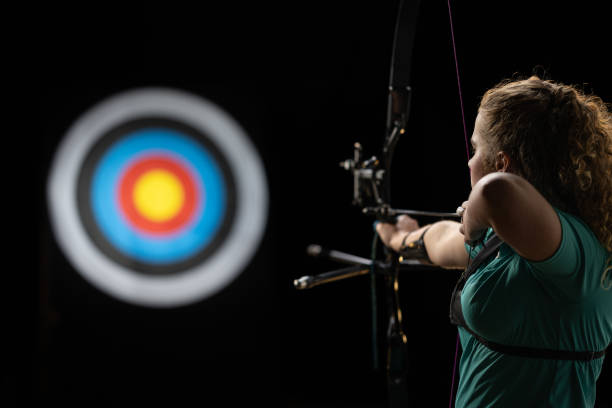 Teenage Caucasian female archer in a studio setting wearing a teal shirt and holding a bow. She is intensely focused on shooting an arrow towards a target illuminated in the background.