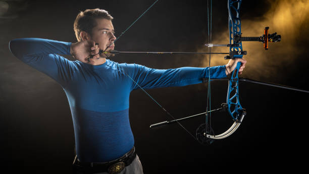 Young Caucasian male archer aiming an arrow with precision in a studio setting, wearing a blue long-sleeve shirt. The setting highlights his athletic skill and concentration in a controlled environment.