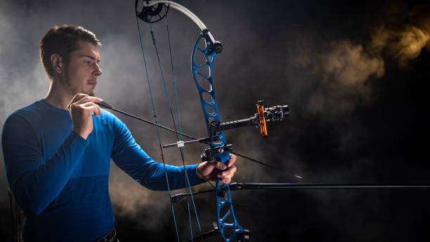 A young Caucasian male archer in a blue shirt carefully places an arrow on a modern compound bow. The scene features dramatic lighting with a smoky background, highlighting the precision and concentration involved in archery.