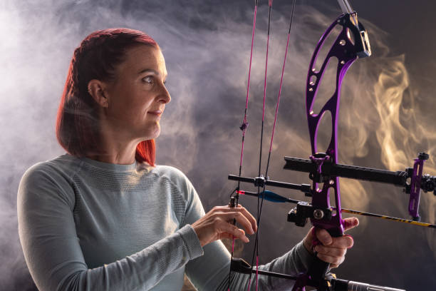 A mid-adult Caucasian woman, wearing a light sweater, skillfully places an arrow on a purple bow. The scene, shrouded in misty smoke, highlights her concentration and expertise in archery, creating a dynamic and engaging depiction.