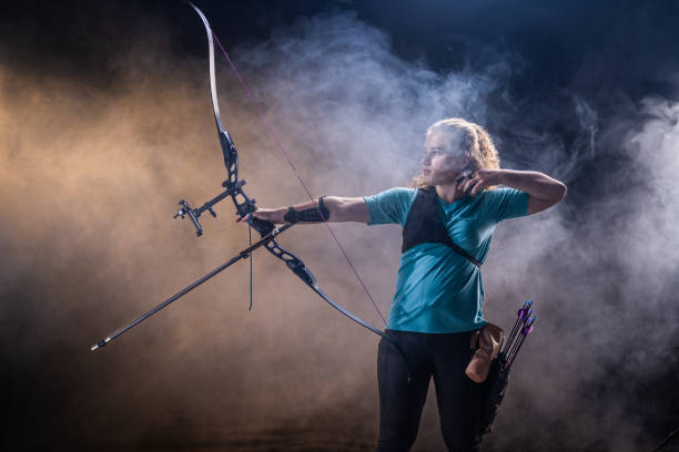 Teenage Caucasian female archer in a teal shirt aiming an arrow in a studio setting with dramatic lighting and smoke, showcasing precision and concentration. Her quiver is strapped to her waist, emphasizing professional archery skills.