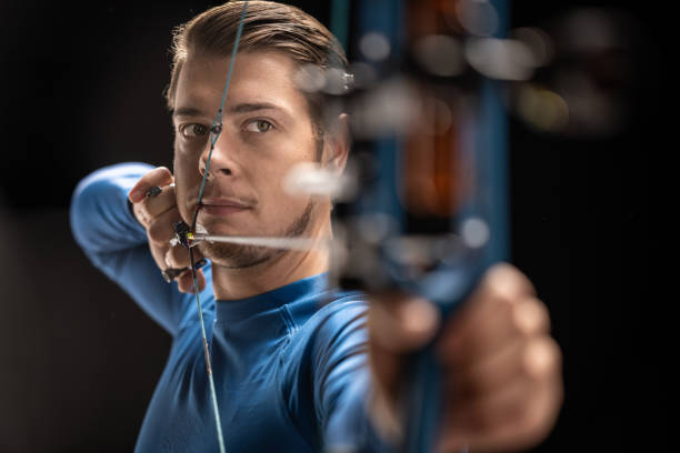 Young adult Caucasian male archer in a studio setting, concentrating as he aims his bow at a target. He wears a blue long-sleeve shirt, highlighting the precision and skill involved in the sport of archery.