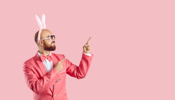 Funny young man wearing easter bunny ears shows on copy space on pastel pink background. Cheerful man in glasses and pink suit advertises or recommends. Creative banner for celebration of Easter.