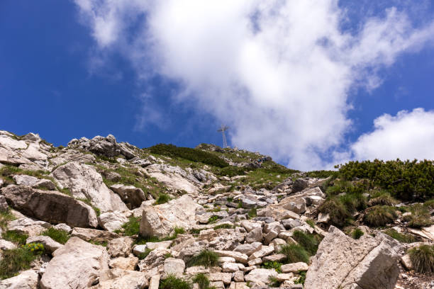 A steep mountain slope with a rocky surface along which a path leads, a cross can be seen on the top, but the sky is covered with white clouds.